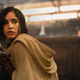 A woman wearing a brown, hooded poncho kneels while holding a blaster gun.